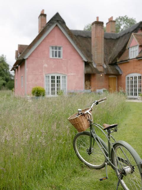Pink thatched cottage behind long meadow grass with a bicycle in the foreground