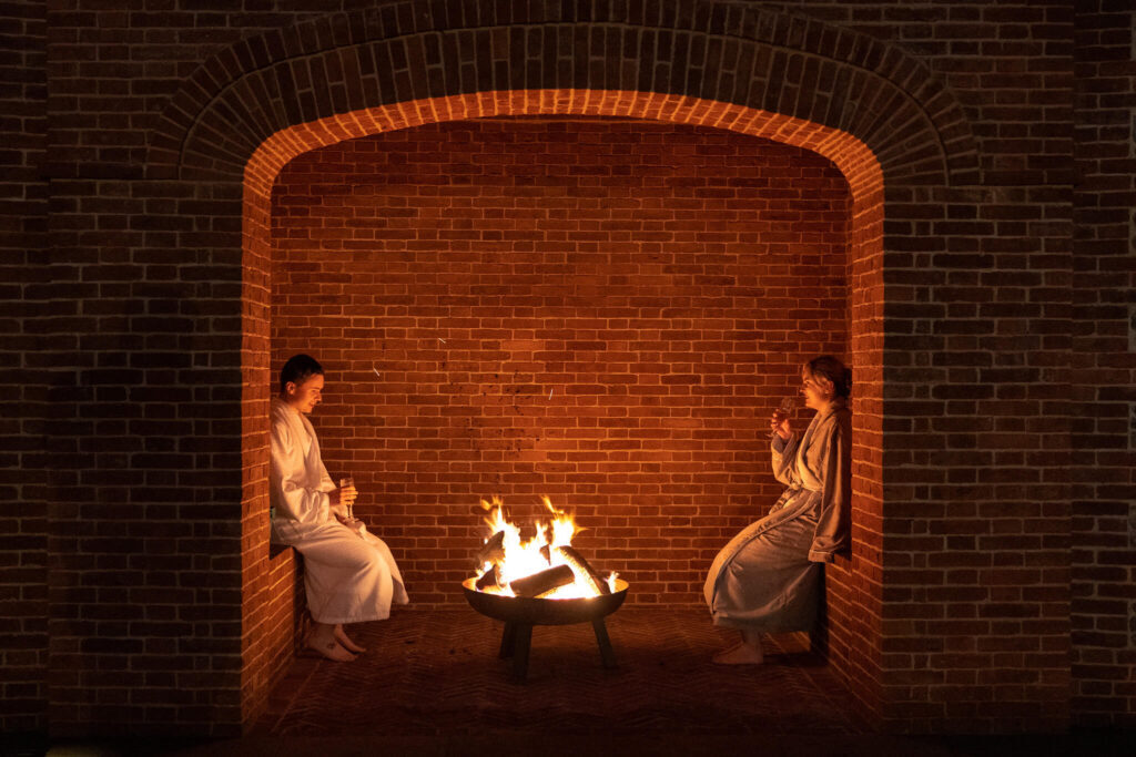 Two guests in robes beside a fire pit within a brick archway at night